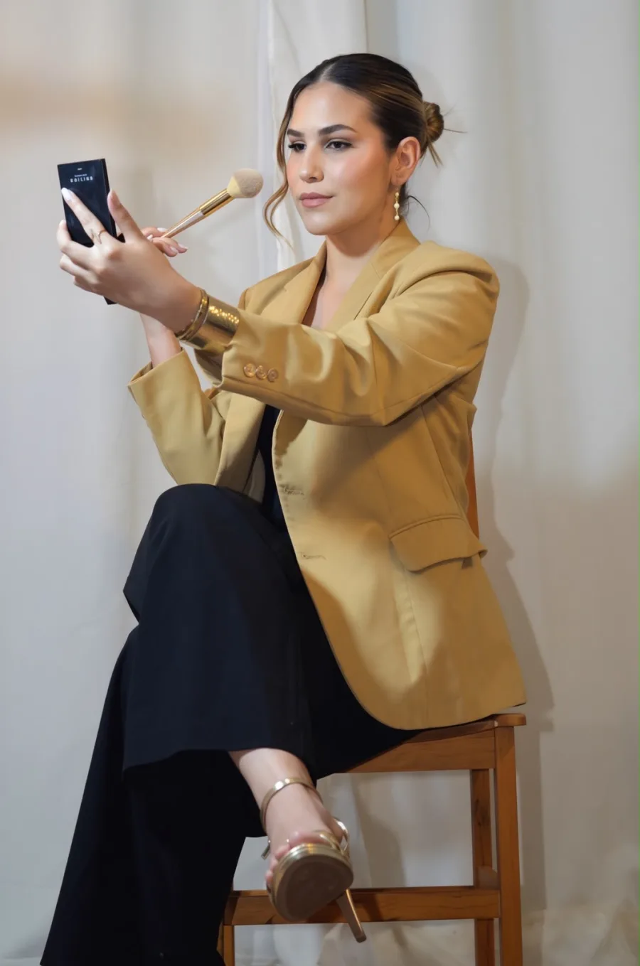 Laura Silveira, maquiadora, de blazer caramelo, retocando a maquiagem com pincel e espelho